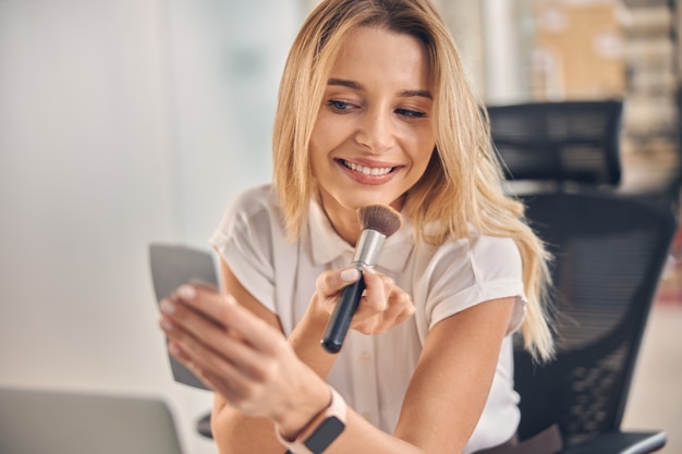 Bella signora bionda che si guarda allo specchio e sorride mentre si trucca in ufficio