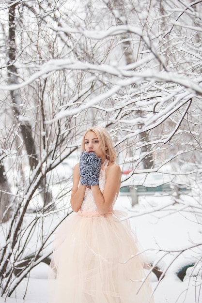 ピンクのドレスと手袋をはめた美しいブロンドは、青い公園の背景に雪を投げます