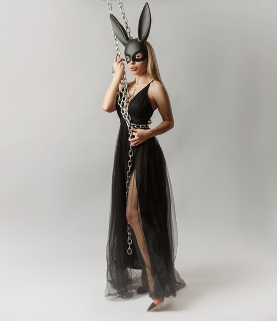 The beautiful blonde in a rabbit mask in a sexy evening dress on a white background with a metal chain