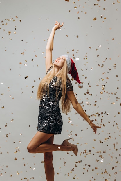 Beautiful blonde Santa girl dancing under glittering confetti New Year party concept