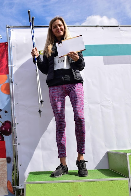 A beautiful blonde sportswoman from Nordic walking on a pedestal shows the won prize and smiles