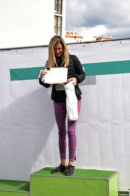 A beautiful blonde sportswoman from Nordic walking on a pedestal shows the won prize and smiles