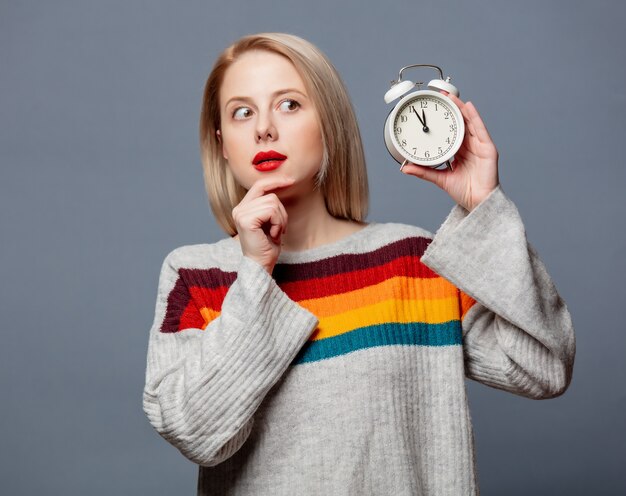 Beautiful blonde in sweater with alarm clock on gray