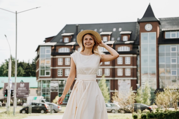 A beautiful blonde walks through a city. Woman in white dress and straw hat