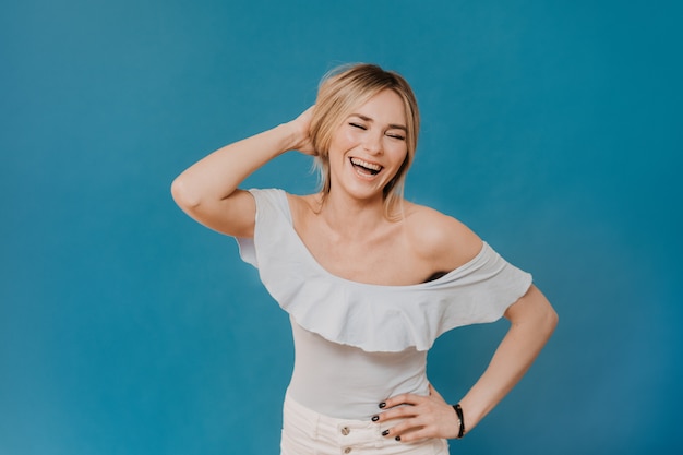 Beautiful blonde wearing Light blouse and white jeans laughing loudly, enjoying free time, preparing for party, over blue background with copy space. Positive people concept.