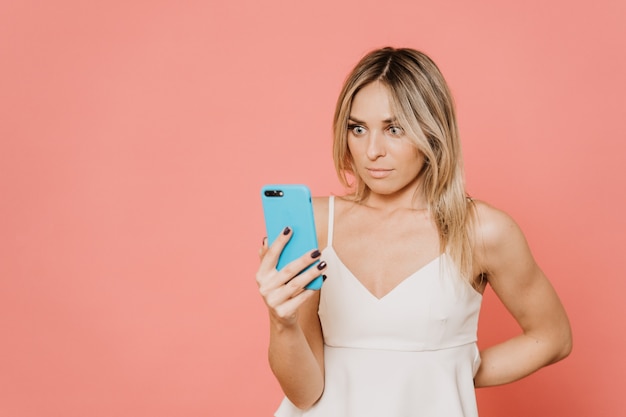 Beautiful blonde in white blouse with wide open eyes staring at her cellphones screen, shocked after reading a message. Over pink backdrop with a copy space. People emotions concept.