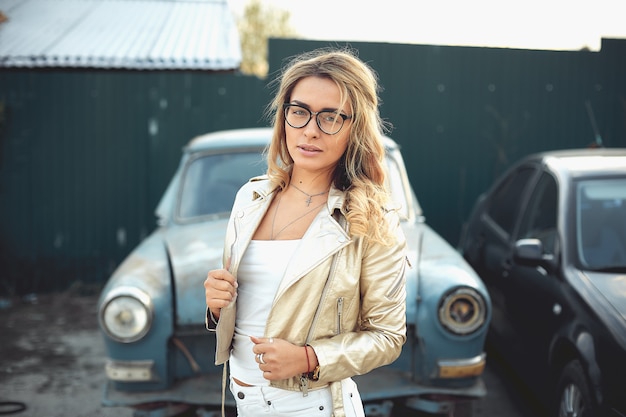 A beautiful blonde with long hair in white denim shorts stands near an old retro car