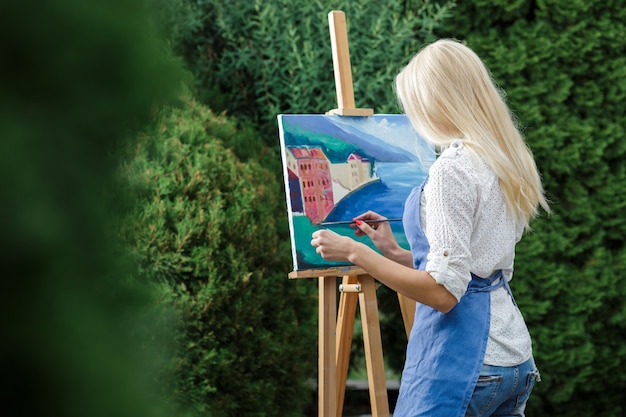 Photo beautiful blonde woman artist with a brush in her hand draws on canvas in the garden.