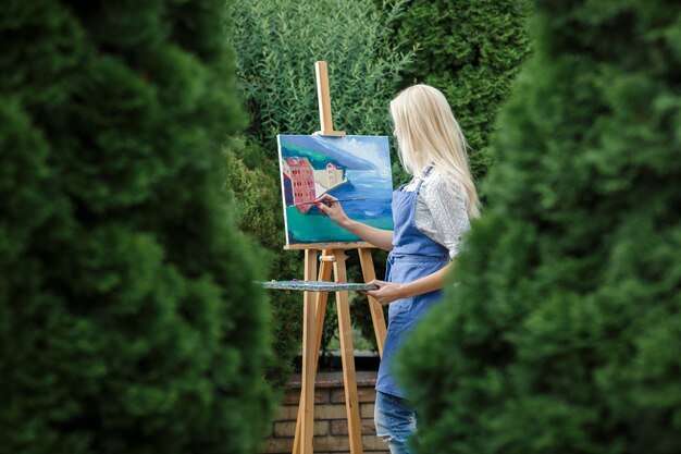 Photo beautiful blonde woman artist with a brush in her hand draws on canvas in the garden.