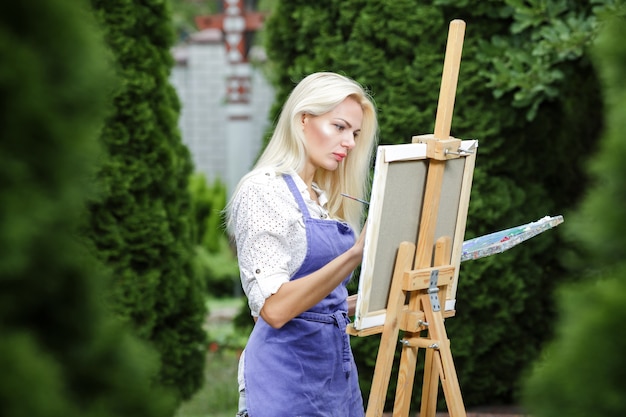 Artista di bella donna bionda con un pennello in mano disegna su tela in giardino.