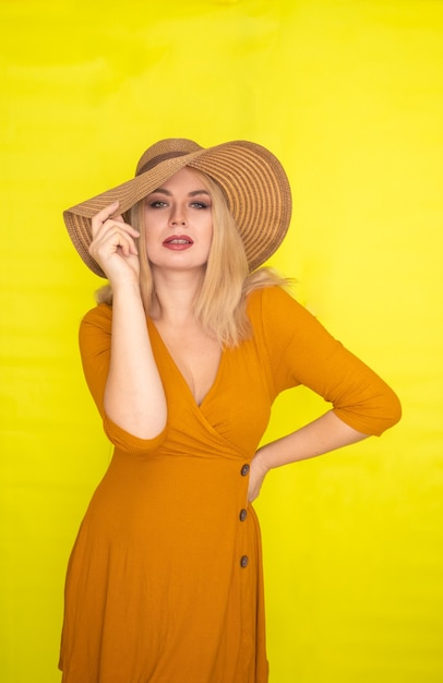 Beautiful blonde woman in brown hat and dark yellow dress posing over yellow wall. Fashion summer look