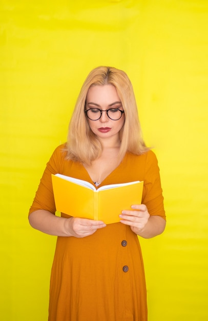 Beautiful blonde woman in glasses holding book in her hands over yellow wall