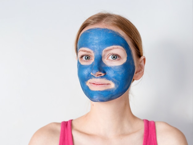 青い粘土の顔のマスクを持つ美しい金髪の女性は、美容師によって適用されます。