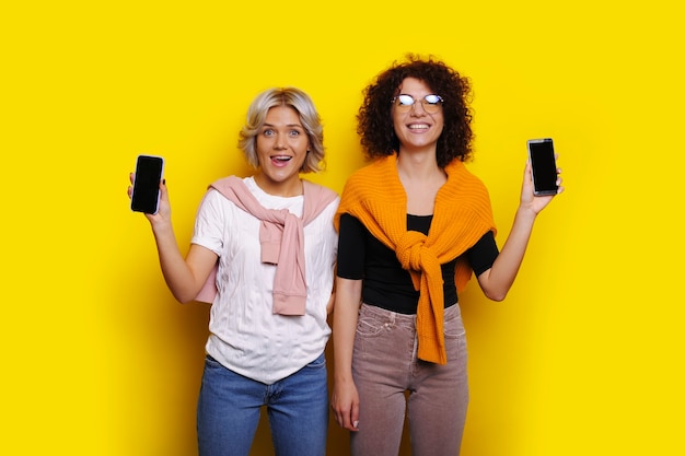 Bella donna bionda e la sua adorabile sorella riccia che guarda l'obbiettivo che ride mentre fa pubblicità al loro smartphone isolato sulla parete gialla dello studio