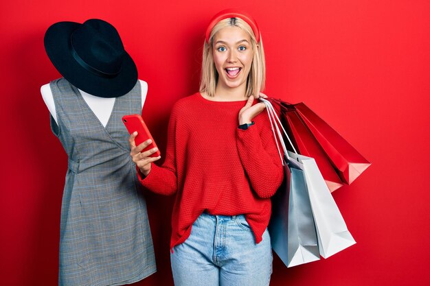 Photo beautiful blonde woman holding shopping bags and smartphone celebrating crazy and amazed for success with open eyes screaming excited