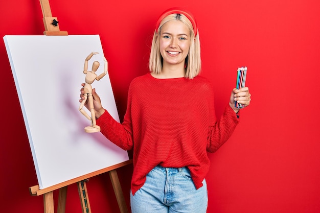 Beautiful blonde woman holding small wooden manikin and pencils smiling with a happy and cool smile on face. showing teeth.