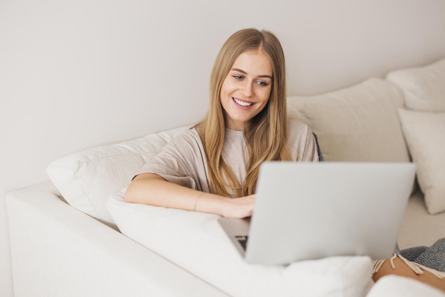 잠옷을 입은 아름다운 금발 여성은 소파에 앉아 집 검역소에서 일하는 노트북 개념을 연구하고 있습니다.