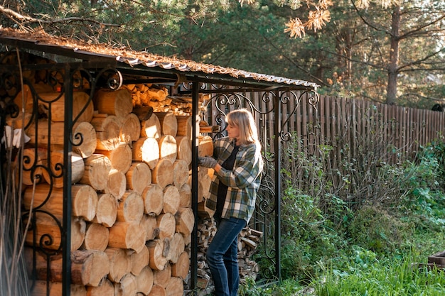 格子縞のシャツを着た美しいブロンドの女性は、ウッドバーナーの日常の田舎の生活から木を収集します