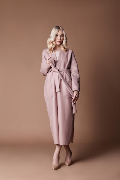 Beautiful blonde woman posing in pink coat on beige background. Fashion show clothing, woman with long hair. Trendy fashionable autumn coat