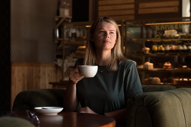 美しいブロンドの女性は喫茶店とパン屋に座って、カプチーノコーヒーを飲みます