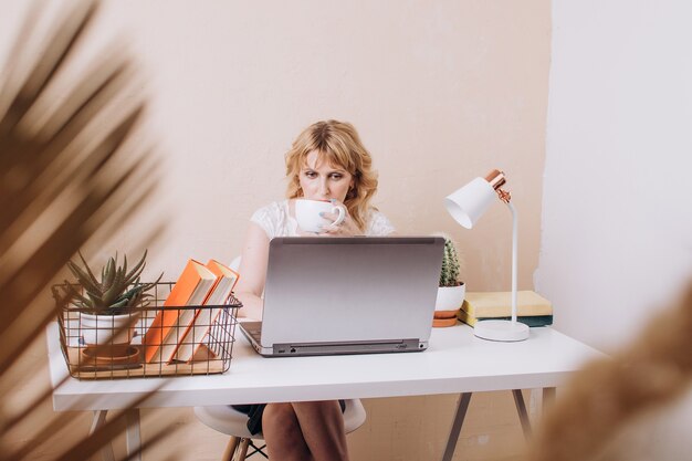 美しいブロンドの女性がラップトップに座って働いて、コーヒーやお茶を飲みます