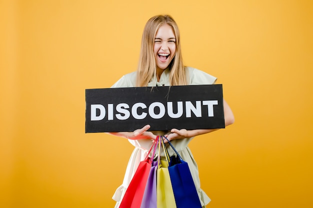 Photo beautiful blonde woman smiling and screaming with discount sign and colorful shopping bags isolated over yellow