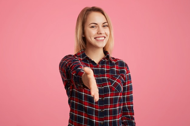 Beautiful blonde woman stretches hand in front, greets somebody, being glad to meet with friend, poses against pink wall. Young female going to shake hands with somebody, gestures indoor