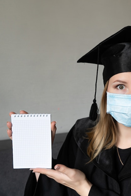 Foto bella donna bionda che indossa il berretto da laurea con maschera facciale e abito da cerimonia con striscione bianco