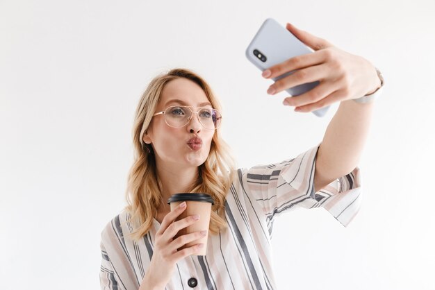 beautiful blonde woman wearing glasses taking selfie photo on smartphone and holding paper cup isolated over white wall