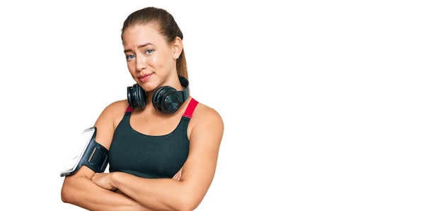 Beautiful blonde woman wearing gym clothes and using headphones looking to the side with arms crossed convinced and confident