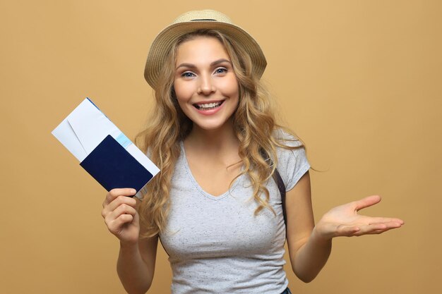 Bella donna bionda che indossa abiti estivi in posa con passaporto con biglietti su sfondo beige.