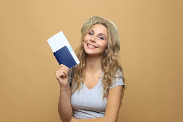 Bella donna bionda che indossa abiti estivi in posa con passaporto con biglietti su sfondo beige.