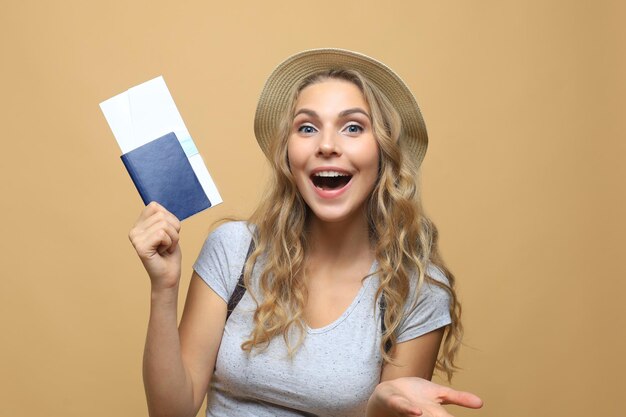 Beautiful blonde woman wearing summer clothes posing with passport with tickets over beige background.