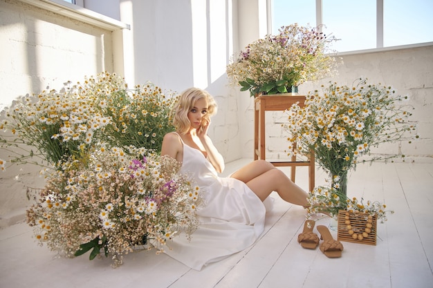 흰 드레스에 아름다운 금발의 여자는 카모마일 꽃 사이에서 바닥에 앉아있다