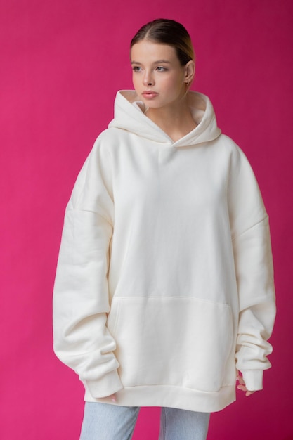 Beautiful blonde woman in a white hoodie and blue jeans posing on a pink background