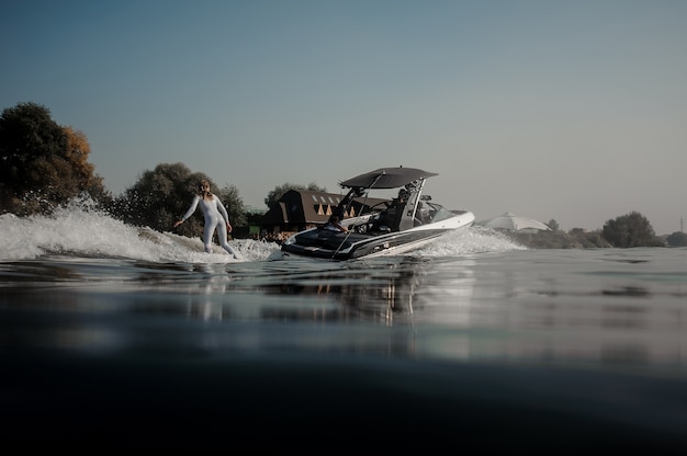 Bella donna bionda in costume da bagno bianco che cavalca sul wakeboard tenendo una corda del motoscafo