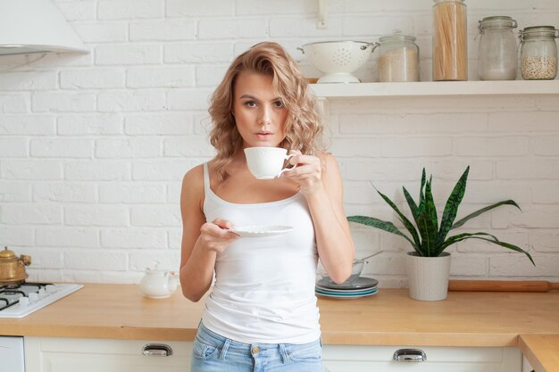 Beautiful blonde woman in white top drinking coffee at home