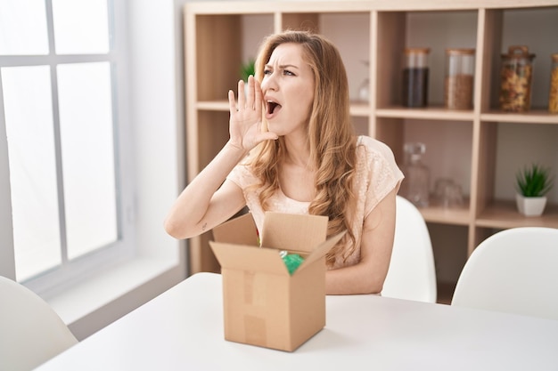 Beautiful blonde woman with cardboard box clueless and confused with open arms, no idea and doubtful face.