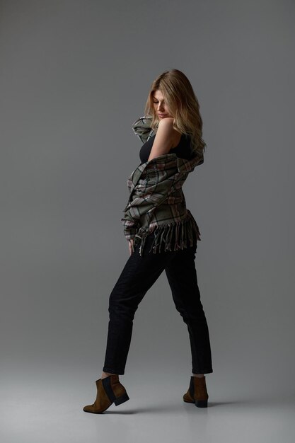 Beautiful blonde woman with curly hair in jacket sitting on chair on studio background