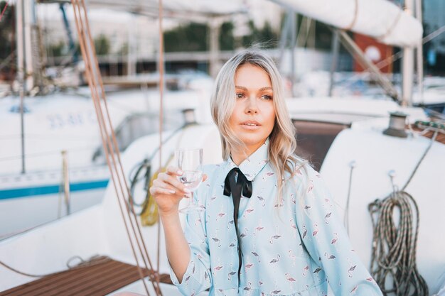 Beautiful blonde young woman in blue dress with glass of soda on boat at pier in the sunset time