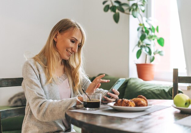 Beautiful blonde young woman having breakfast and using mobile in living room at the home