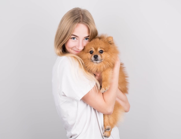 Una bella giovane donna bionda abbraccia e bacia il suo cucciolo di spitz. amore tra cane e padrone. isolato su uno sfondo grigio.