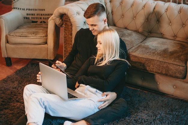 Beautiful blonde young woman leaning on her mans chest sitting on the floor looking to a laptop while man is looking to a tabled smiling indoor.