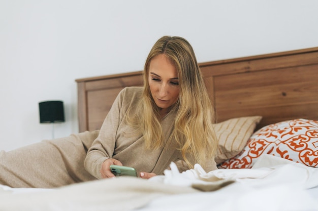 Foto bella giovane bionda che usa il cellulare a letto in una stanza luminosa a casa