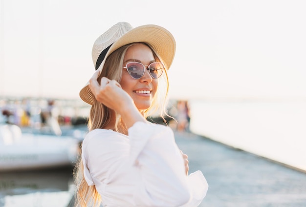 Beautiful blonde young woman in white shirt and straw hat on the pier on sunset