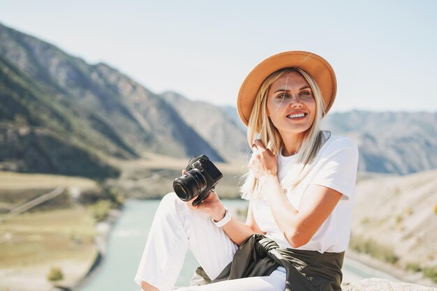 사진 카메라와 함께 펠트 모자를 쓴 아름다운 금발의 젊은 여성 여행자가 알타이 산으로 여행을 갑니다.