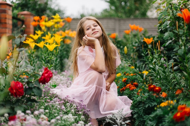 写真 長いブロンドの髪を持つ美しい青い目の少女。ピンクのフラミンゴのドレスの少女。フラワーガーデンの女の子。夏の明るく感動的な写真。家の近くの大きく、厚く、明るいフラワーガーデン。