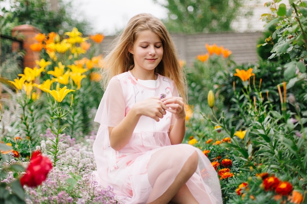 Bella ragazza dagli occhi blu con lunghi capelli biondi. bambina in un vestito di fenicottero rosa. ragazza nel giardino fiorito. estate luminosa, foto emotiva. giardino fiorito grande, spesso e luminoso vicino alla casa.