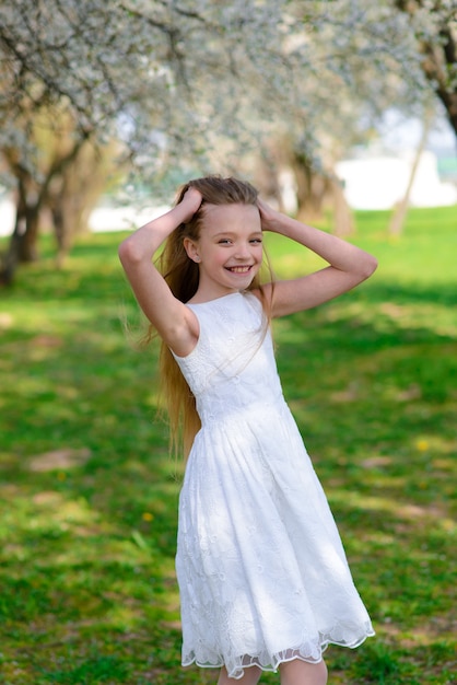 Beautiful blue-eyed girl with long blond hair in a white dress walking in the flower garden. Summer and spring bright, emotional photo.