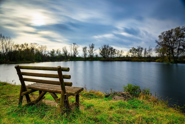 Beautiful blue lake with the trees and the cloudy sky in background and a bench on the shore